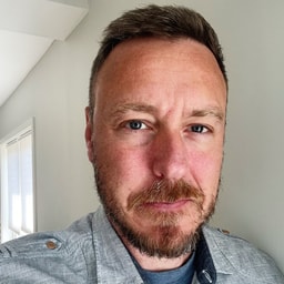 Man Wearing Gray Shirt Standing Indoors Near Window Light Man with short hair and beard wearing a gray shirt standing indoors near window.