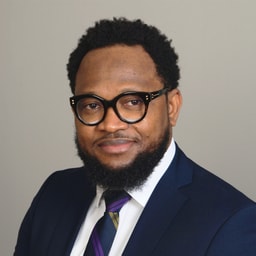 Obinna in navy suit, white shirt, and striped tie against a plain background.