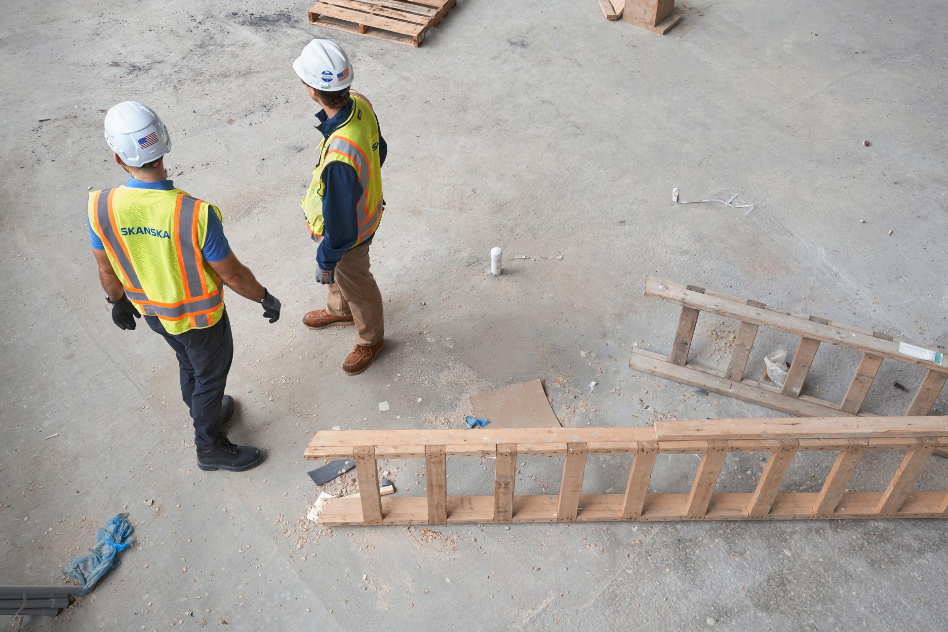 Two construction workers in safety gear discussing plans on a concrete floor site.