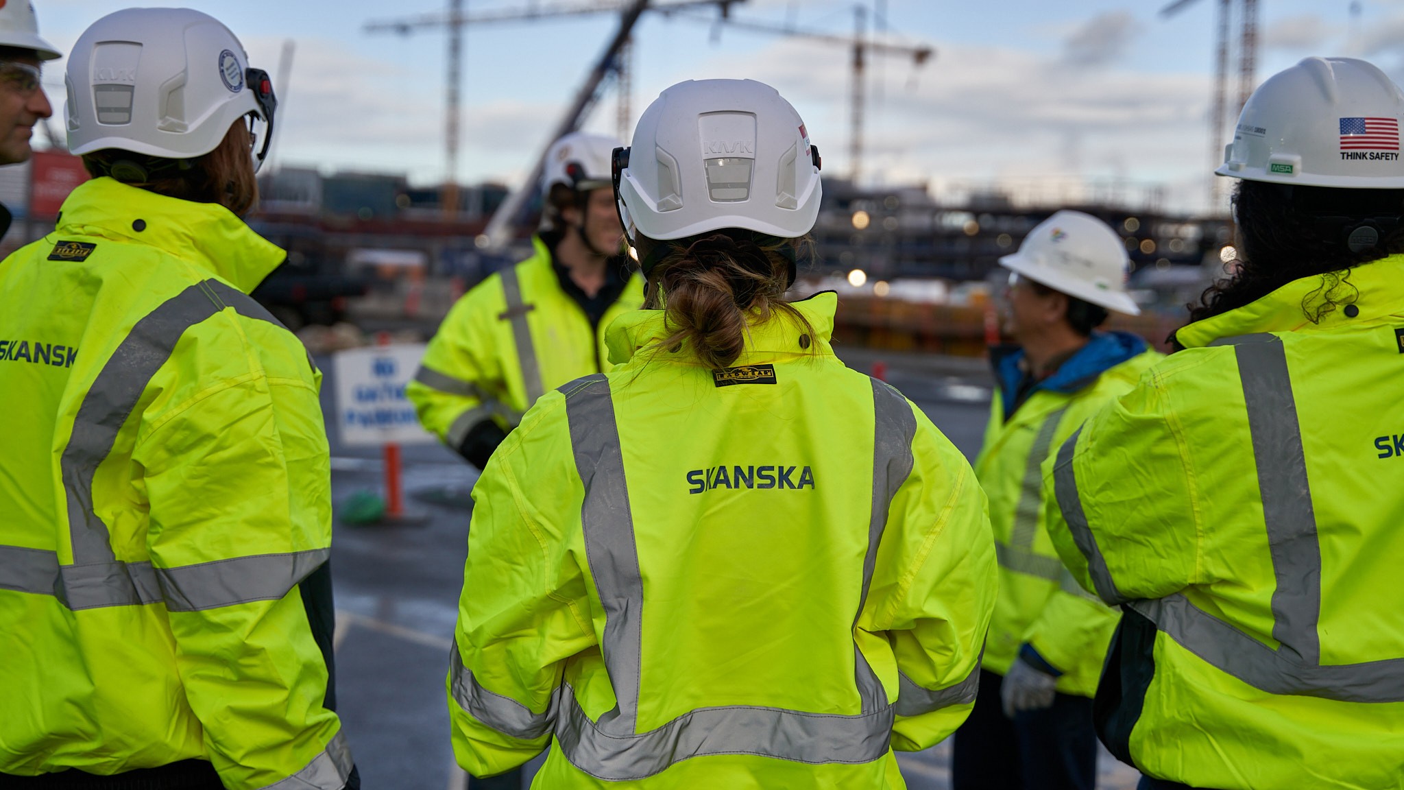 A person in a safety vest stands in front of a construction site with Skanska signage visible.
