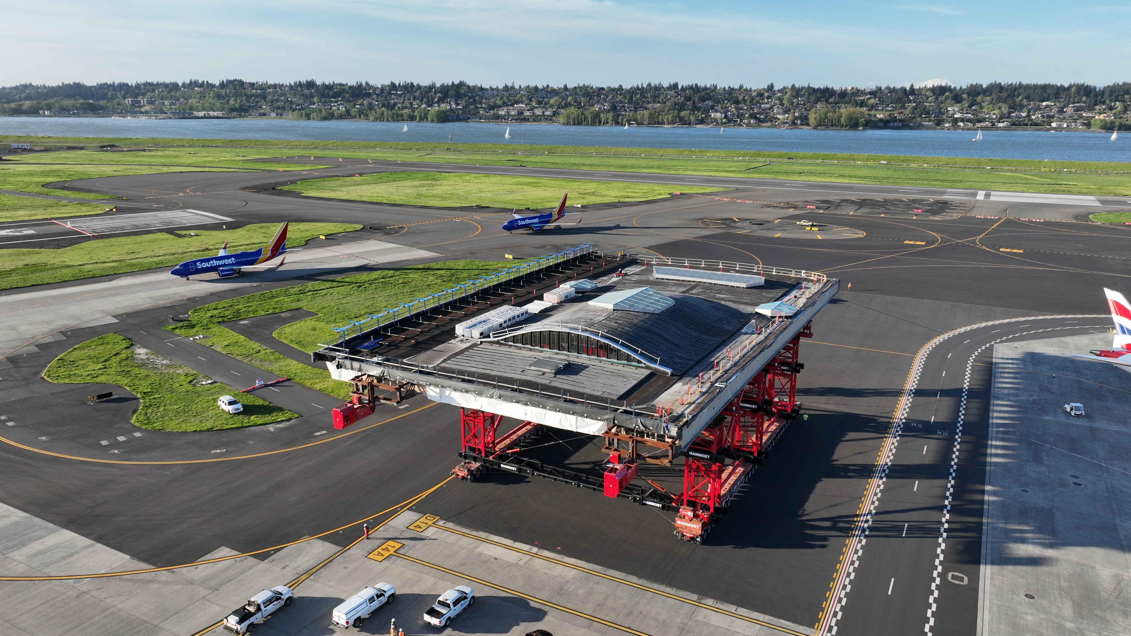 Large structure being transported on red wheeled platforms at an airport, with planes nearby.