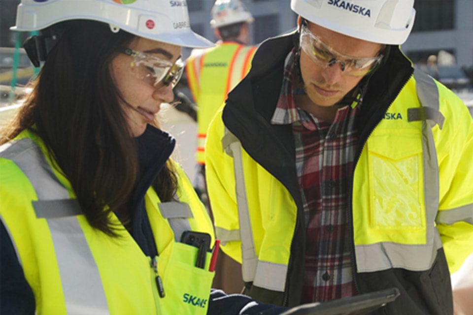 Two construction workers in high-visibility jackets and helmets reviewing plans on site.