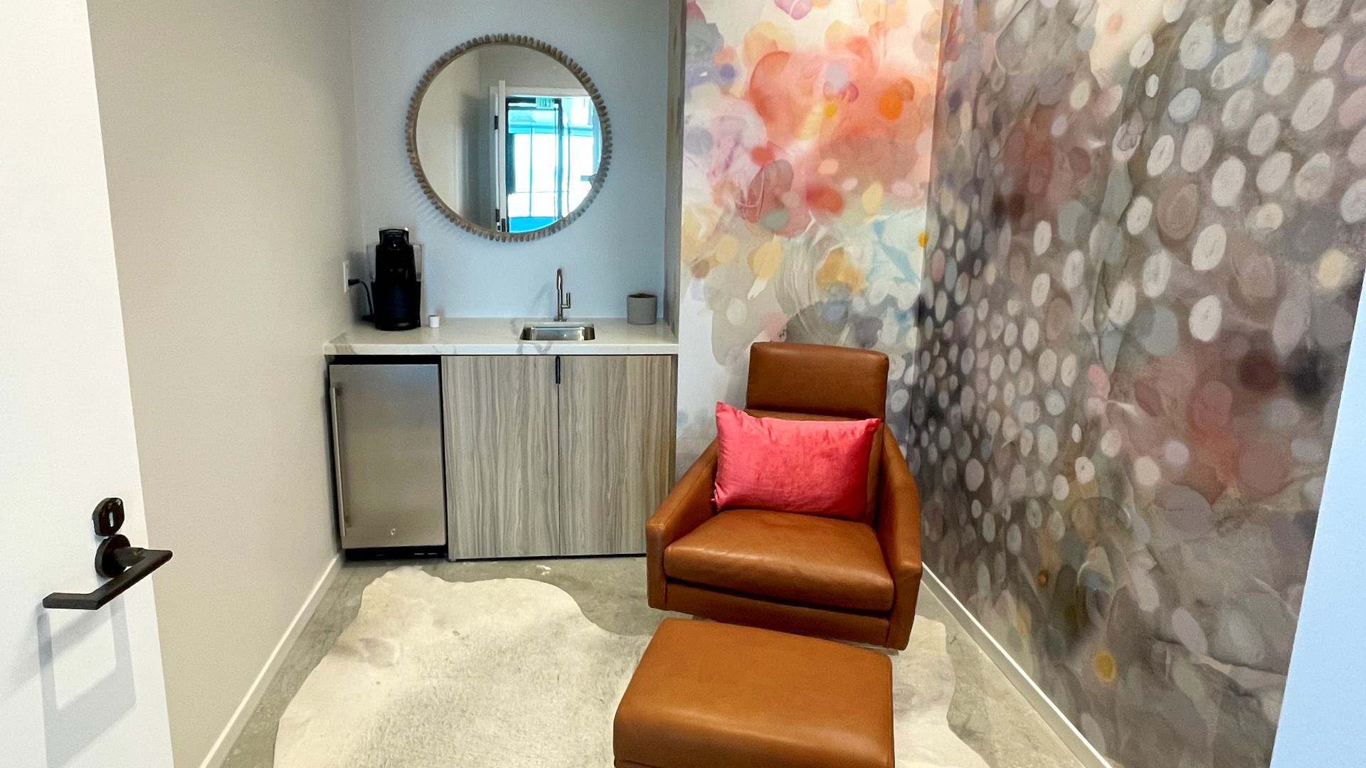 Cozy modern lounge with leather chair, ottoman, colorful wall art, and kitchenette.