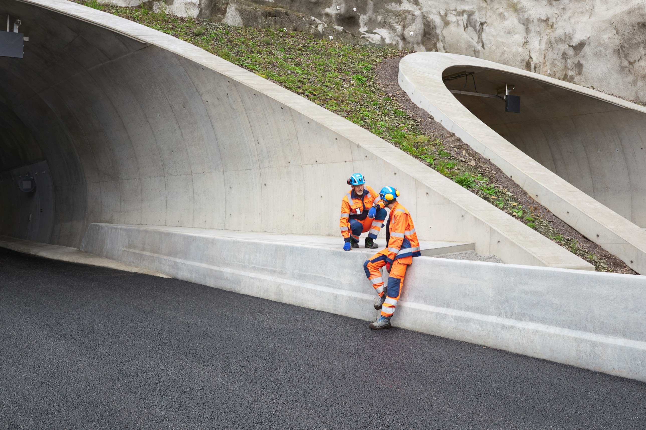Construction workers inspecting modern tunnel entrance site To anleggsarbeidere i oransje sikkerhetsutstyr hviler utenfor en betongtunnelinngang.