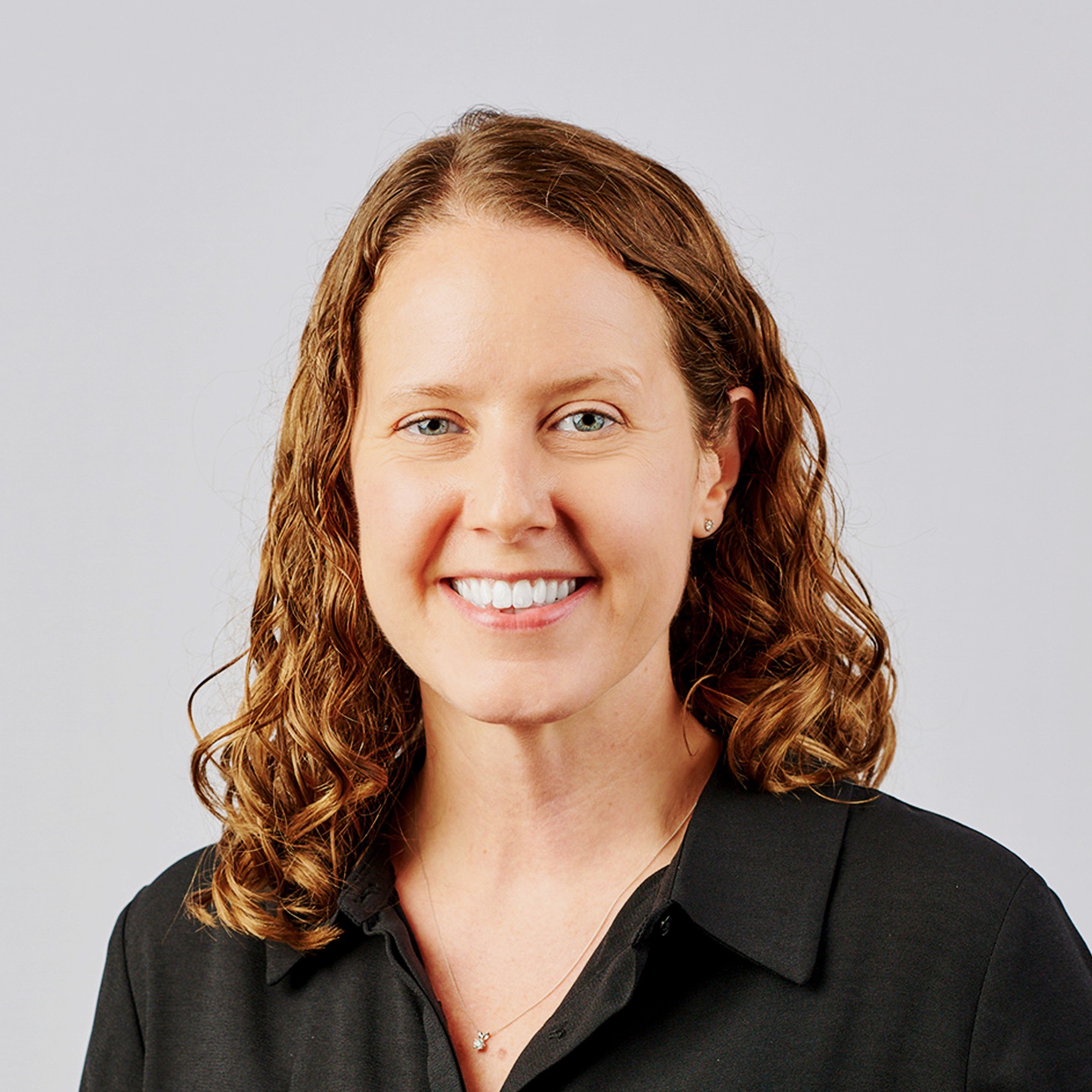 Professional woman with curly hair wearing black shirt and necklace Person with shoulder-length curly brown hair wearing a black collared shirt and necklace.