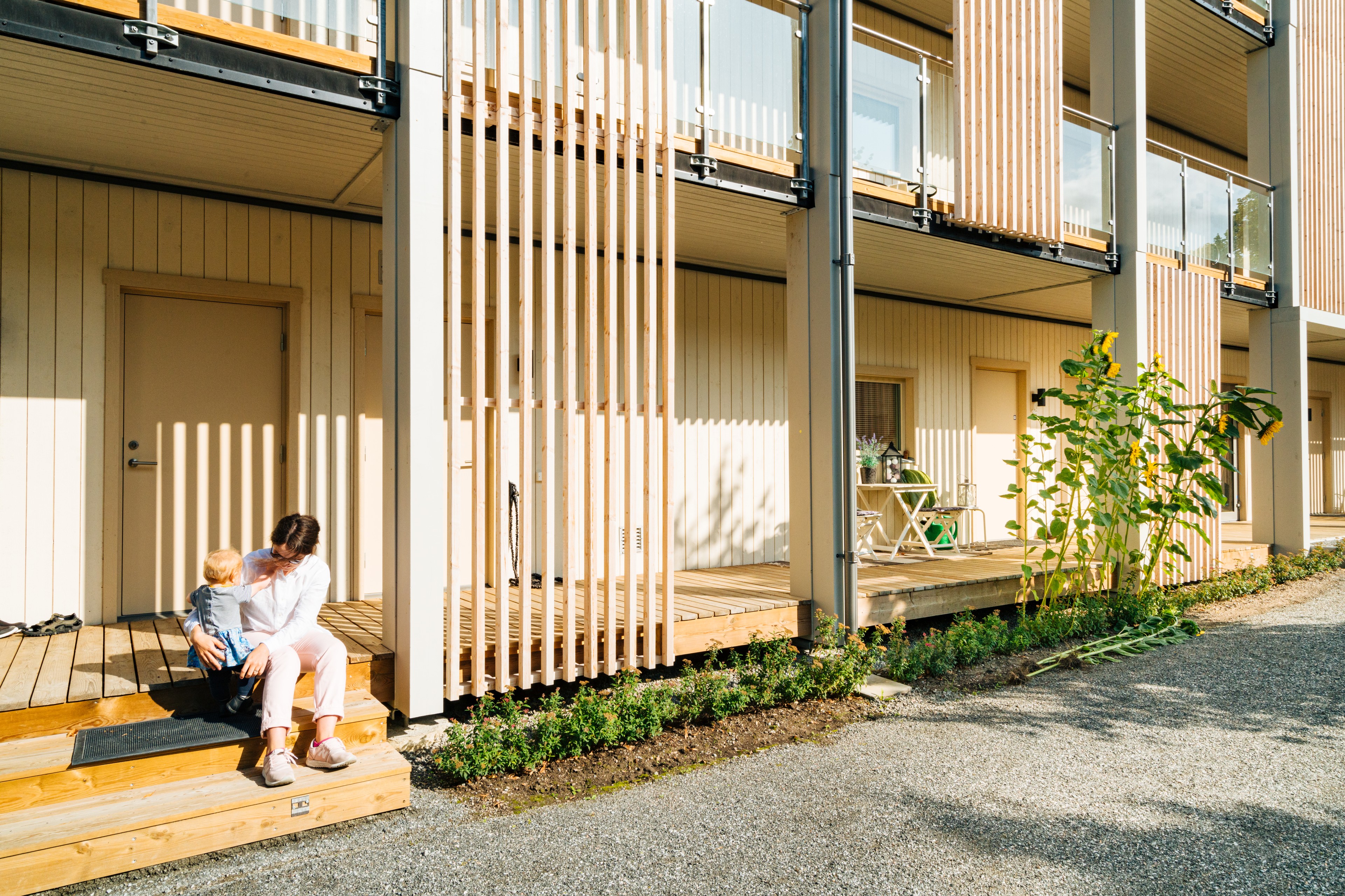 En kvinna sitter på trappsteg i trä med ett barn utanför moderna beige radhus i solsken.