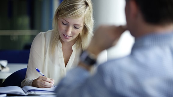 Two people engaged in a business meeting, one taking notes, in an office setting.