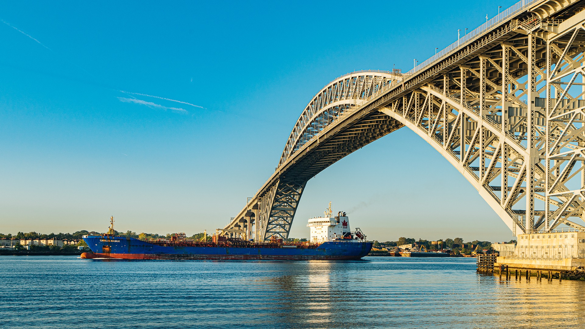 Cargo Ship Passing Under Large Steel Arch Bridge at Sunset Cargo ship passing under a large steel arch bridge over calm blue water at sunset.
