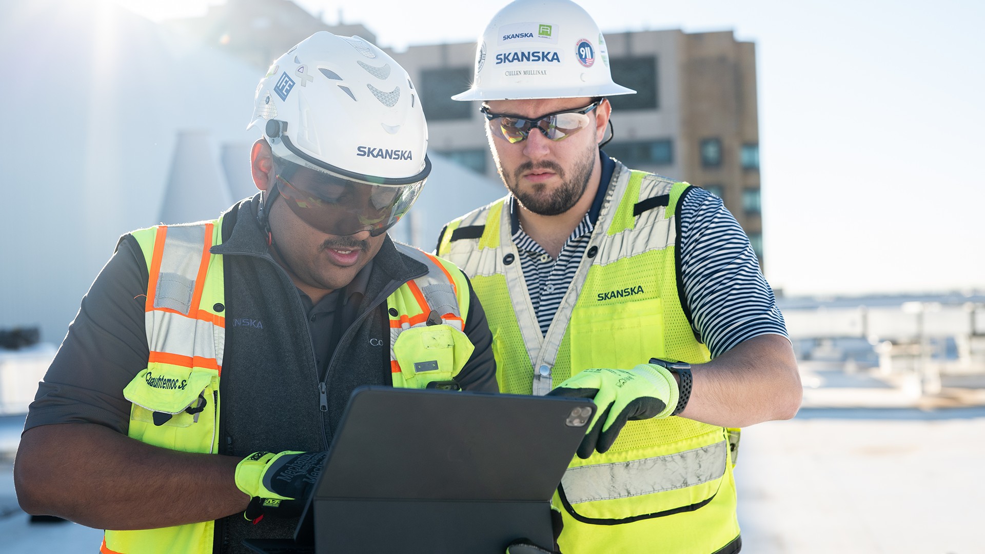 Construction Workers Reviewing Plans on Site Two construction workers in safety gear review a tablet on a sunny outdoor site.