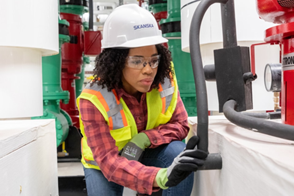 Worker in safety gear operating valve in industrial facility with pipes and gauges.