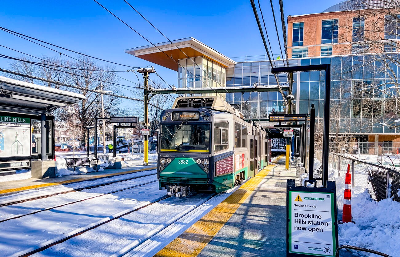 A green trolley at Brookline Hills station on a snowy day, with modern buildings in the background.