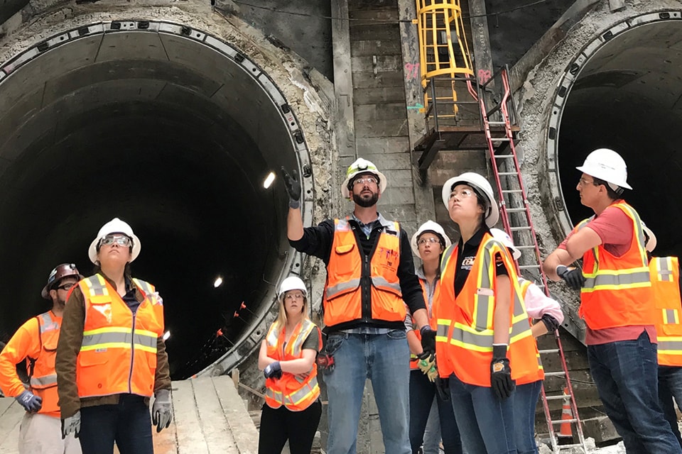 Construction workers and interns in safety gear stand near tunnel entrances at an underground site.
