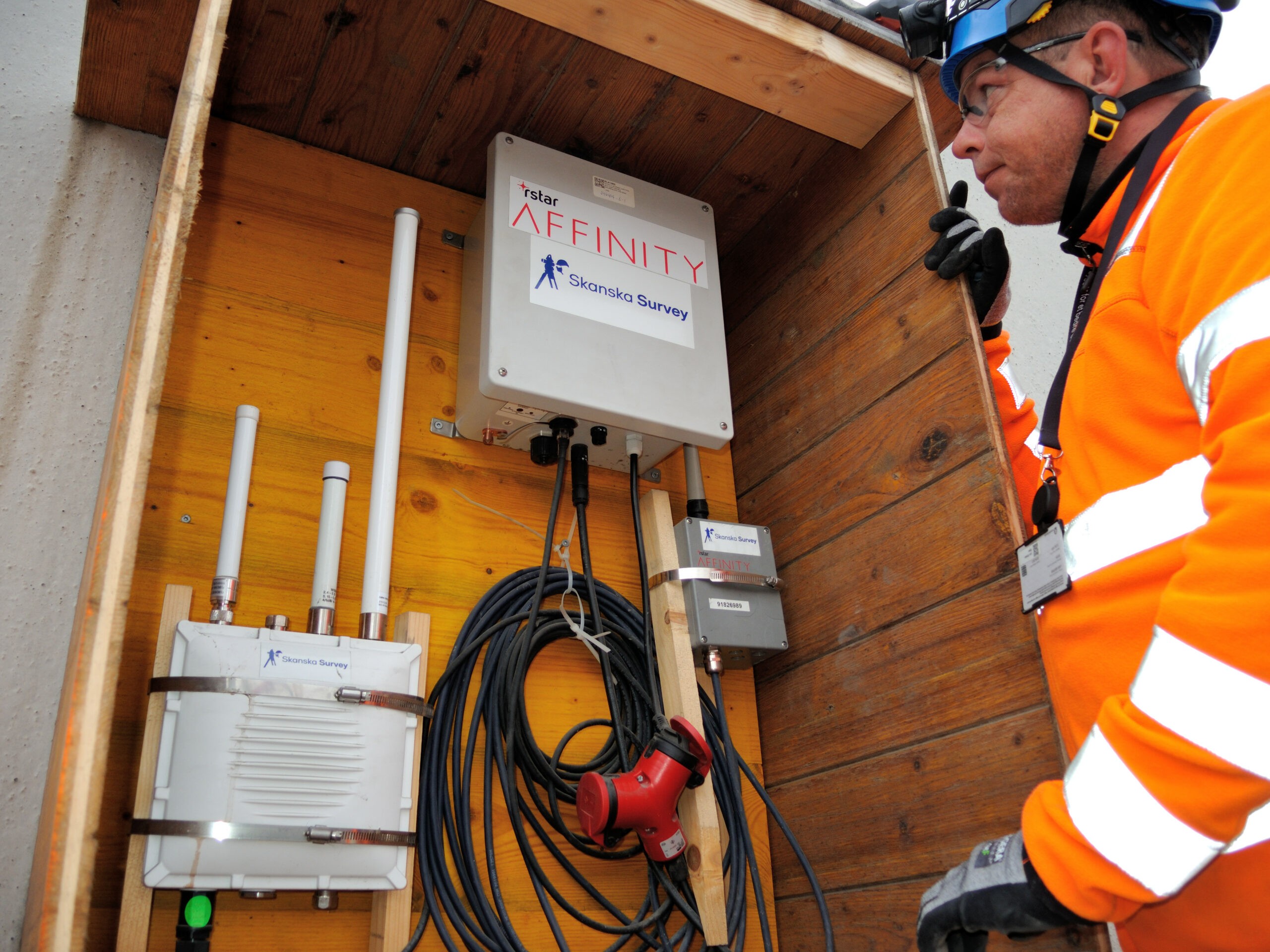Telecommunication Equipment Installation with Safety Worker Bygningsarbeider undersøker elektronisk utstyr merket "AFFINITY Skanska Survey" i en innhegning av tre.
