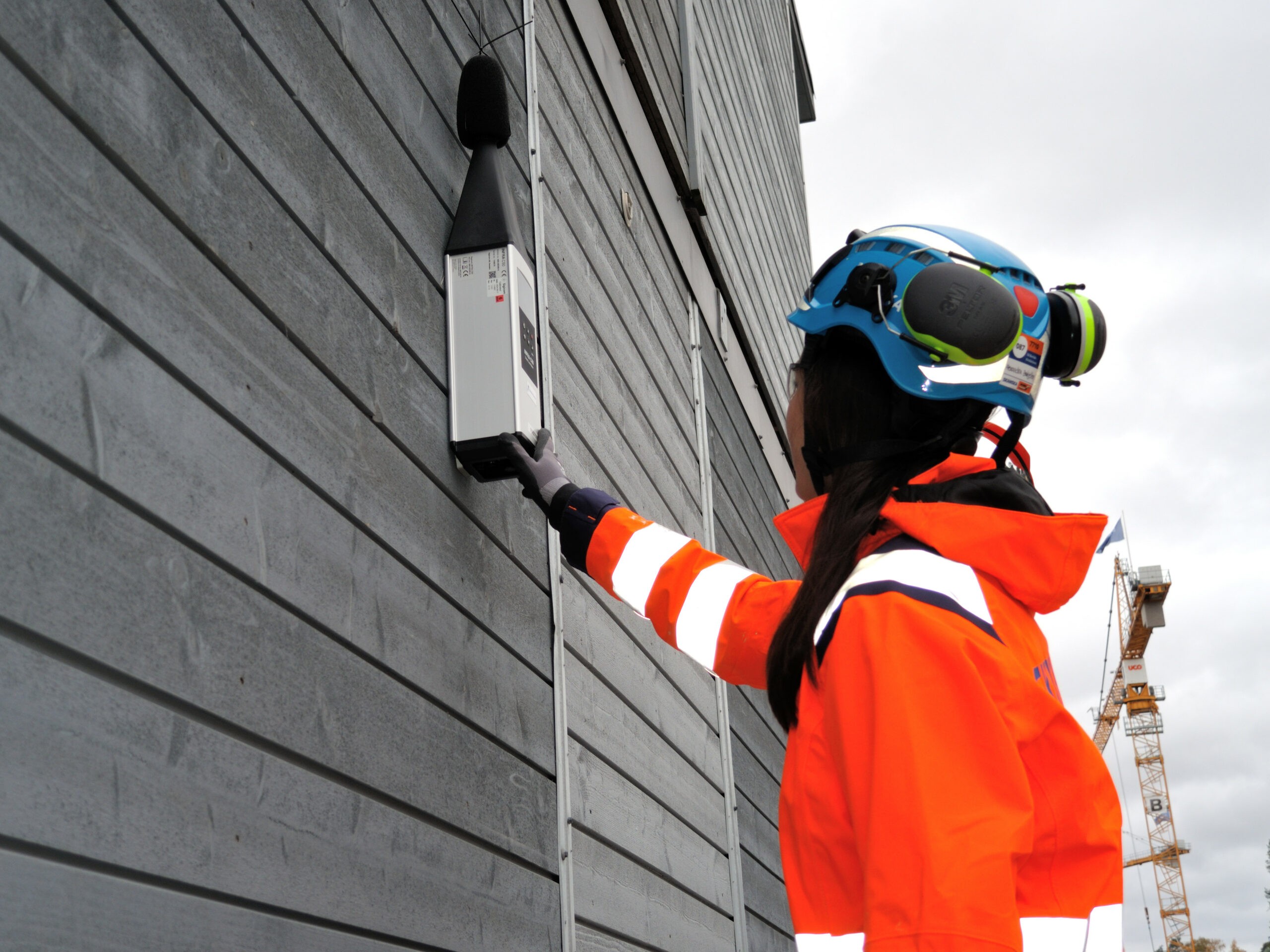 Worker in safety gear inspecting noise monitoring device outdoors En arbeider i verneutstyr installerer en støymåler på en bygningsvegg på en byggeplass.