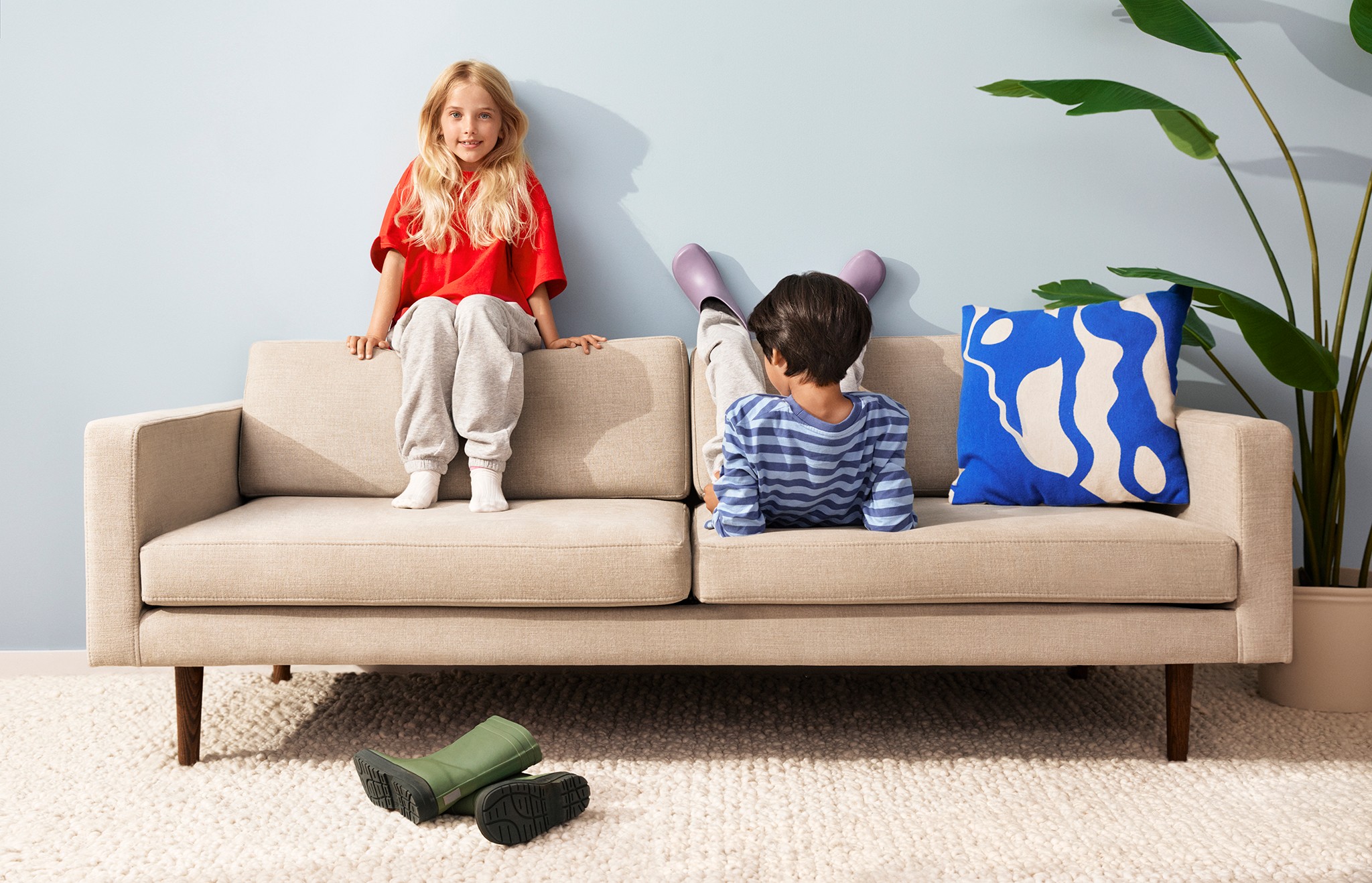 To barn sitter på en beige sofa med en fargerik pute; grønne støvler ligger på teppet i nærheten.