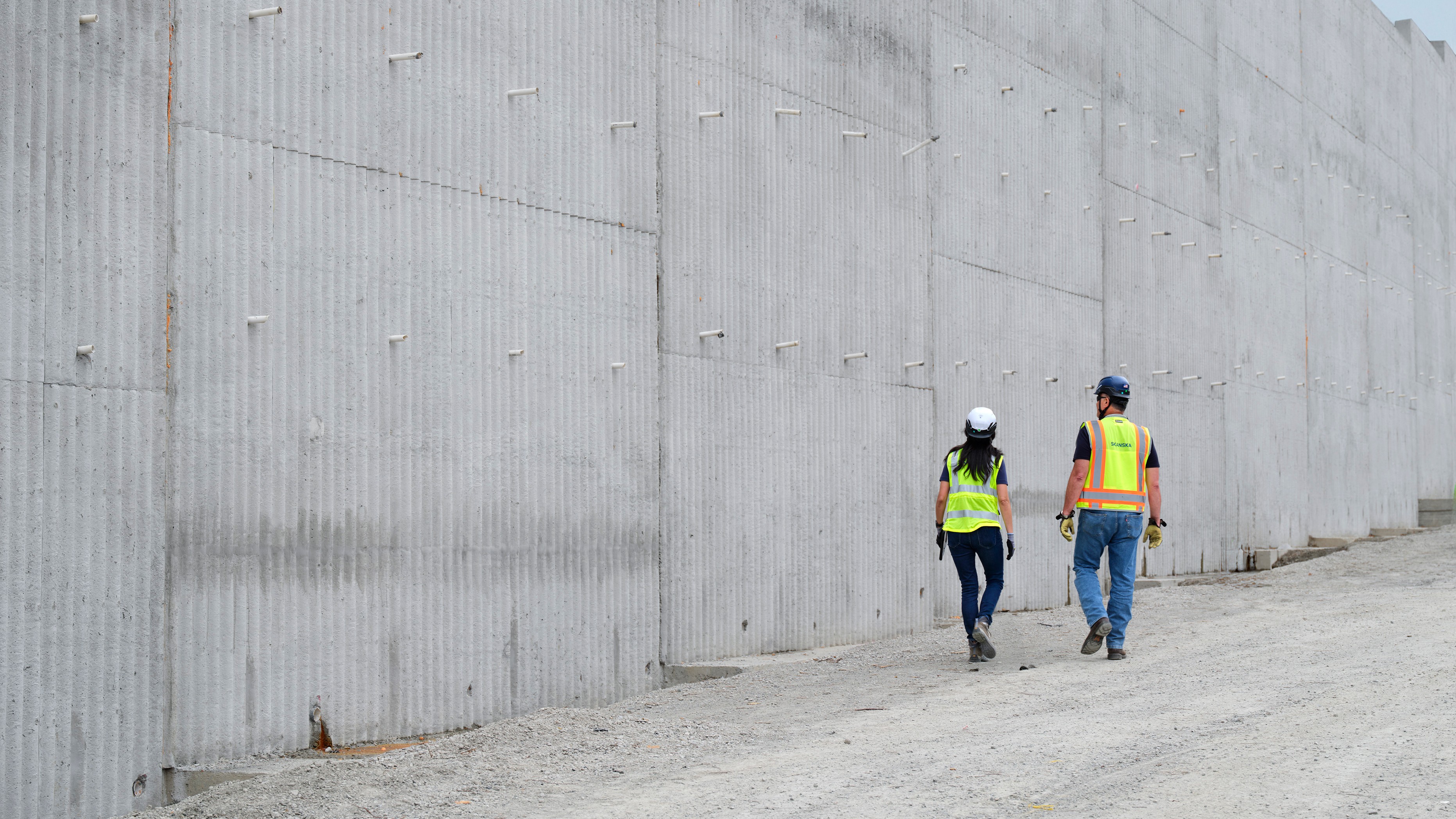 Two construction workers in safety vests walking beside a large concrete wall on site.