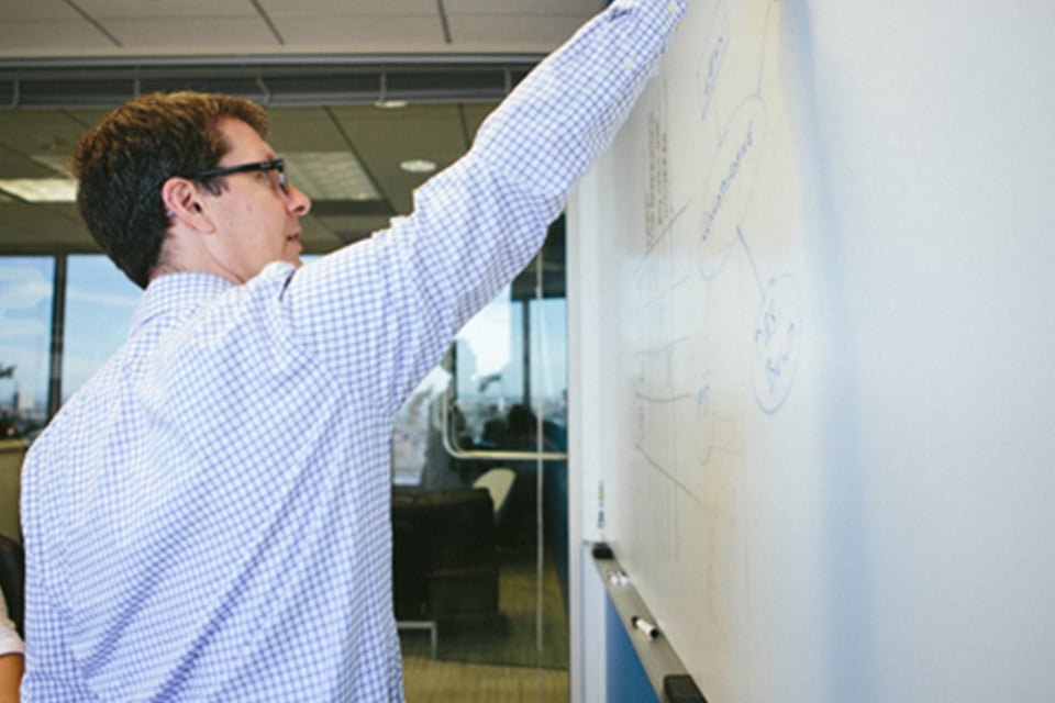 Man in checkered shirt writing on whiteboard in an office setting.