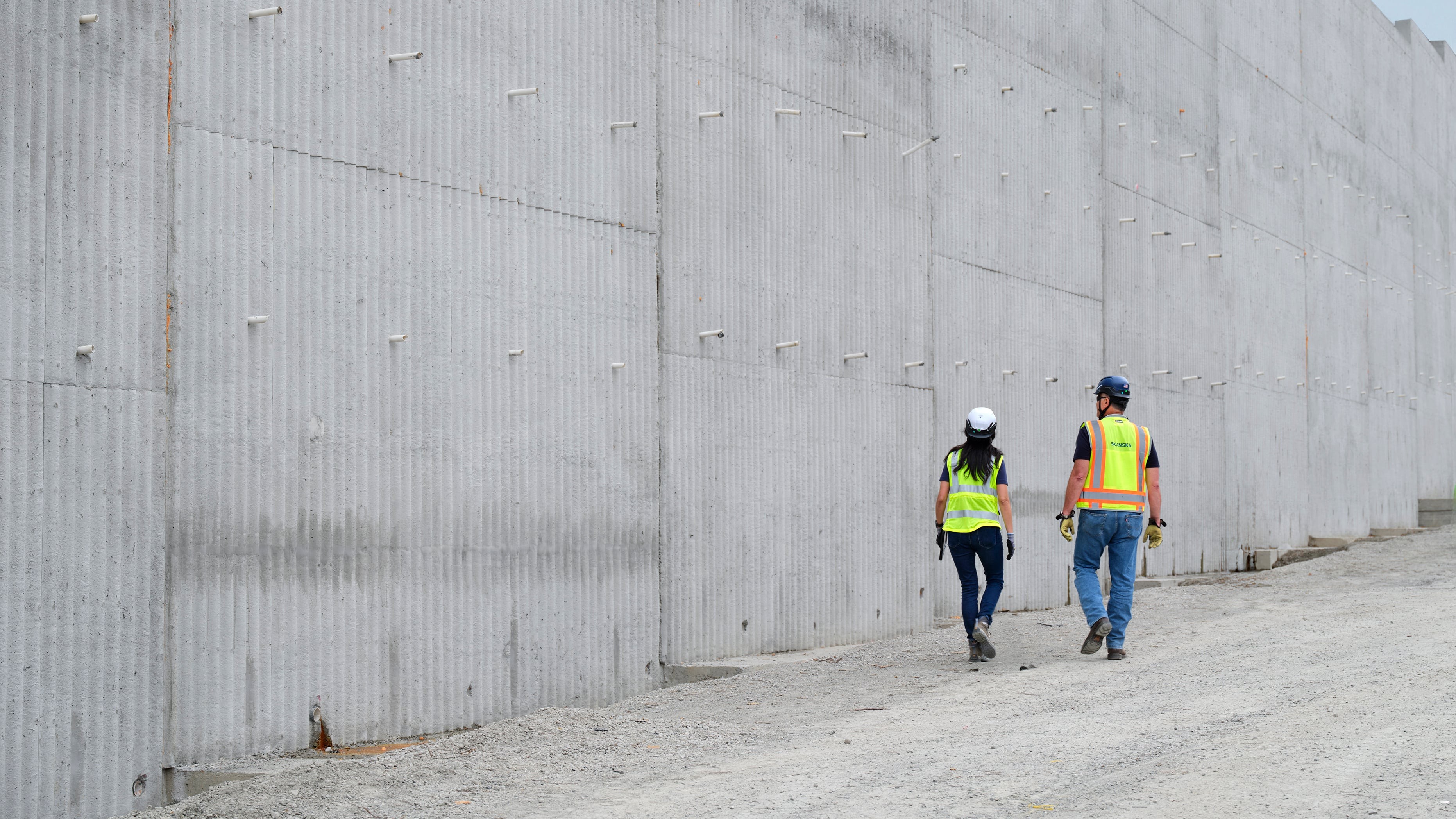 Two construction workers in safety gear walking beside a large concrete wall outdoors.