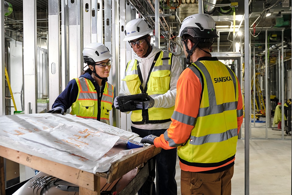 Construction workers in safety gear review plans on-site, wearing Skanska-branded vests.