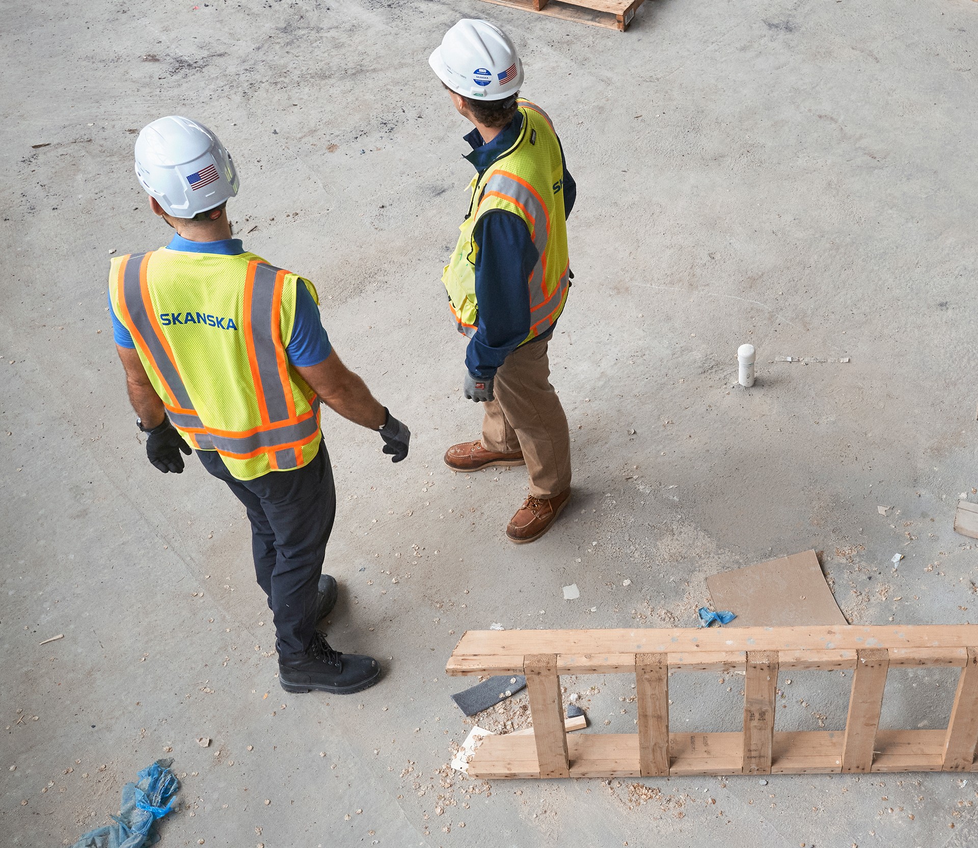 Construction workers in safety gear discussing project plan. Two construction workers in safety vests and helmets discussing on a concrete floor.