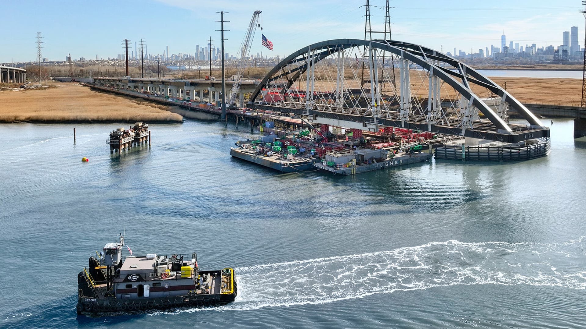 Tugboat pulling a barge with construction materials under an arched bridge over water.