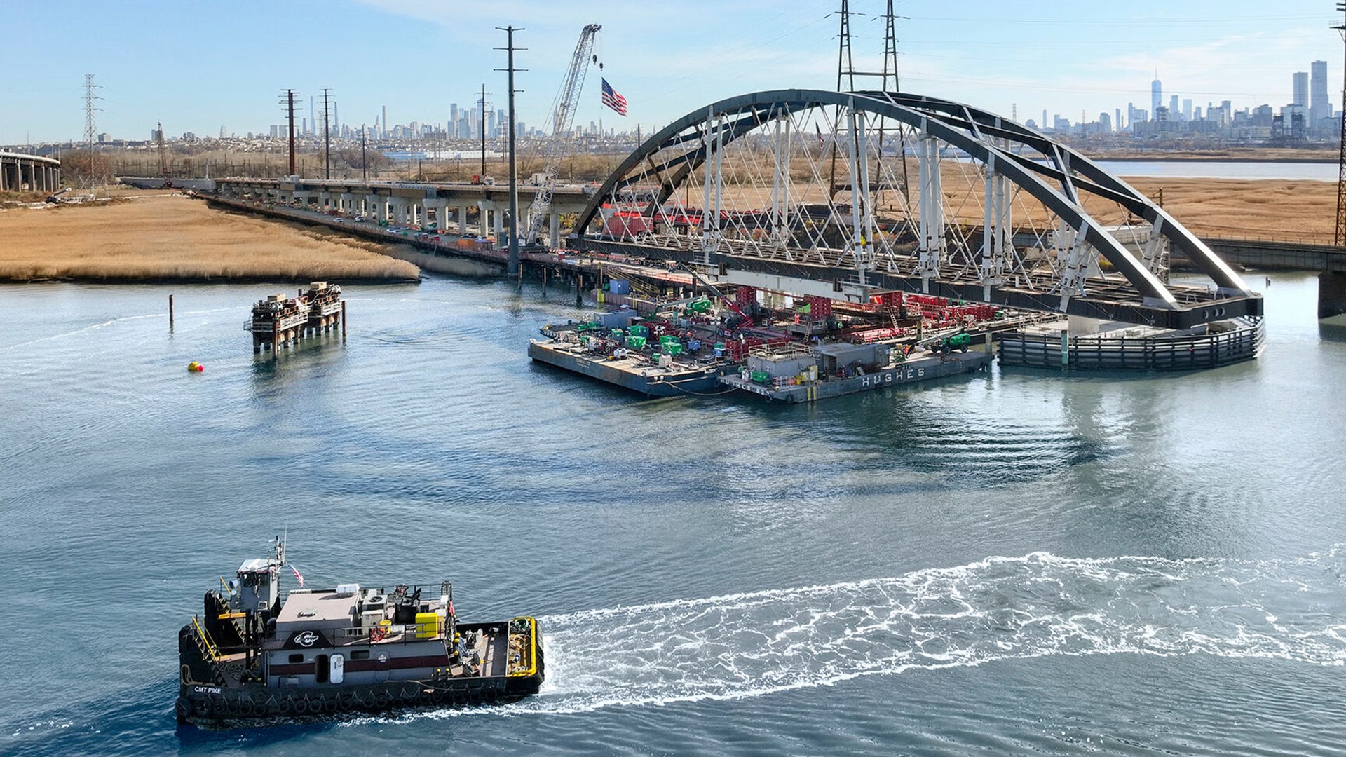 Tugboat pulling a barge with construction materials under an arched bridge over water.