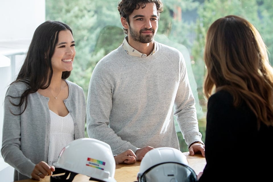 Three professionals discussing plans with safety helmets on a table in a bright office.