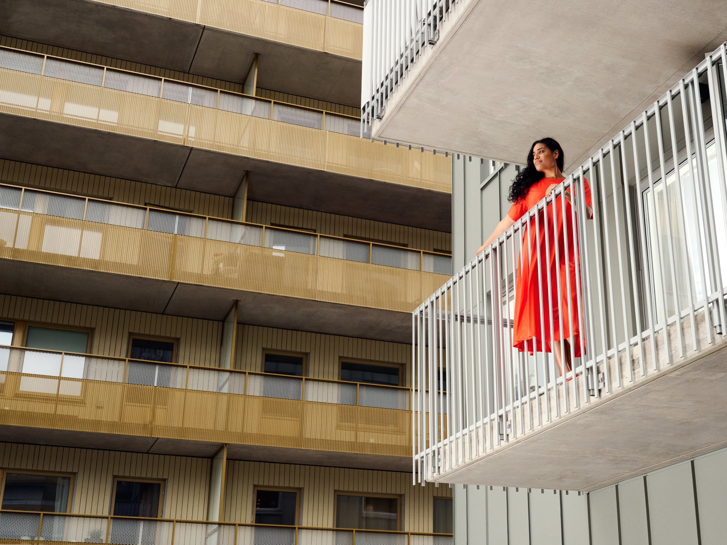 A person in a red dress stands on a balcony of a modern apartment building with geometric design.