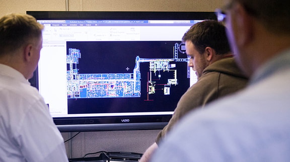 Three people analyze a complex architectural blueprint on a large monitor in an office setting.