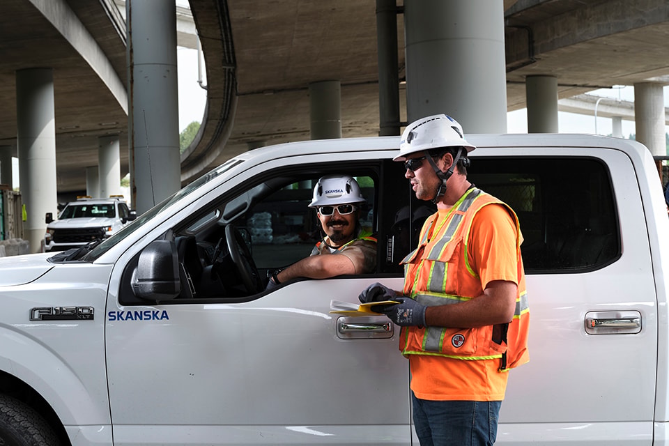 Construction workers in safety gear converse by a Skanska truck under an overpass.