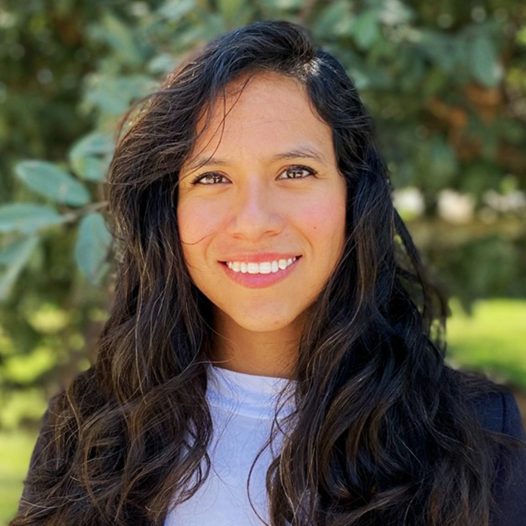 Woman with Long Wavy Hair Outdoors in Natural Green Setting Woman with long wavy hair wearing a white top and dark jacket outdoors near trees.