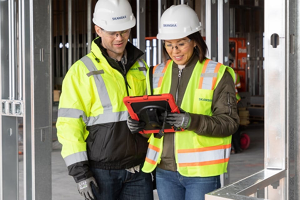 Two construction workers in safety gear using a tablet inside a building site.
