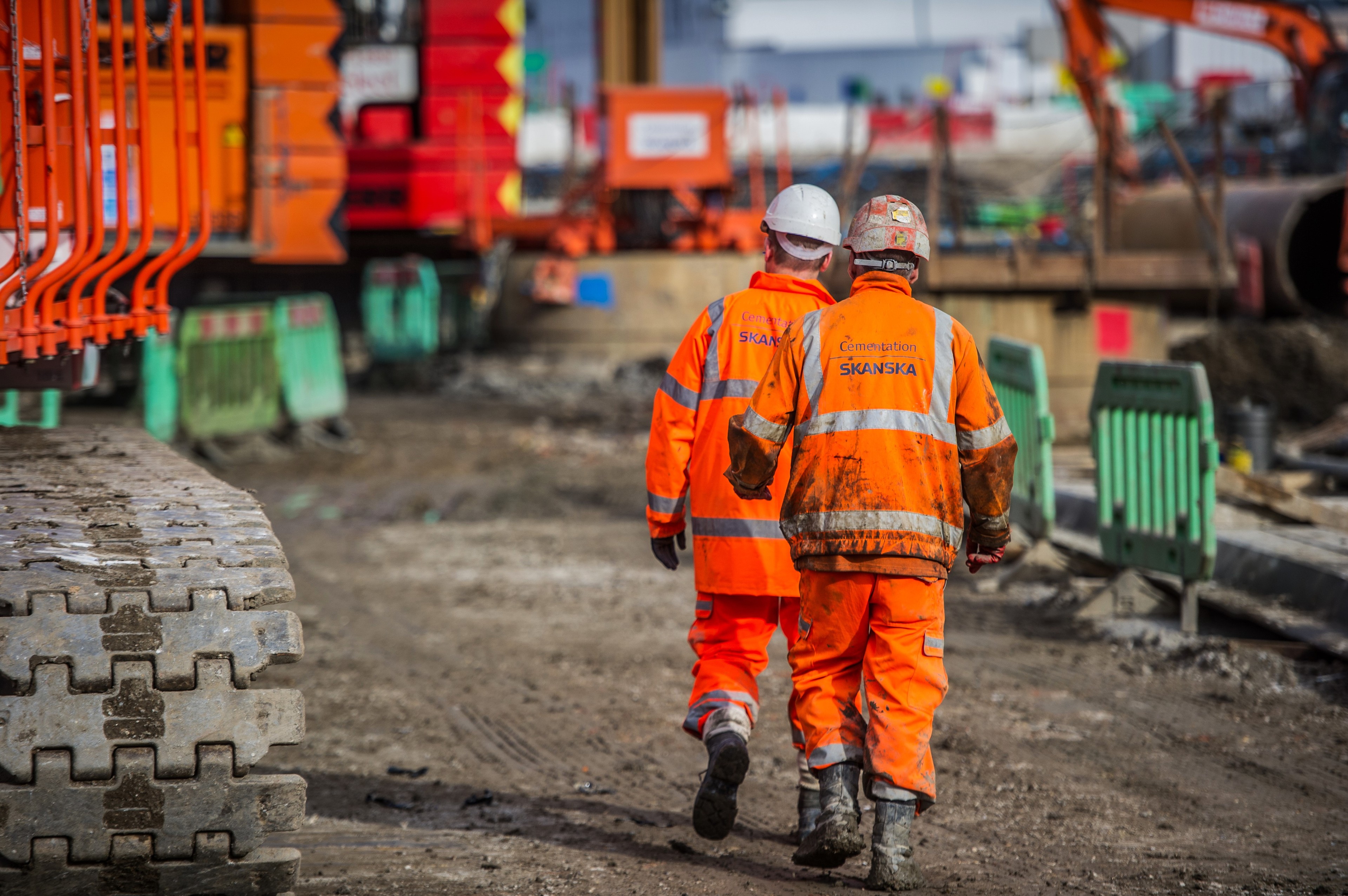 Two construction workers in orange safety gear walking on a muddy site with machinery.