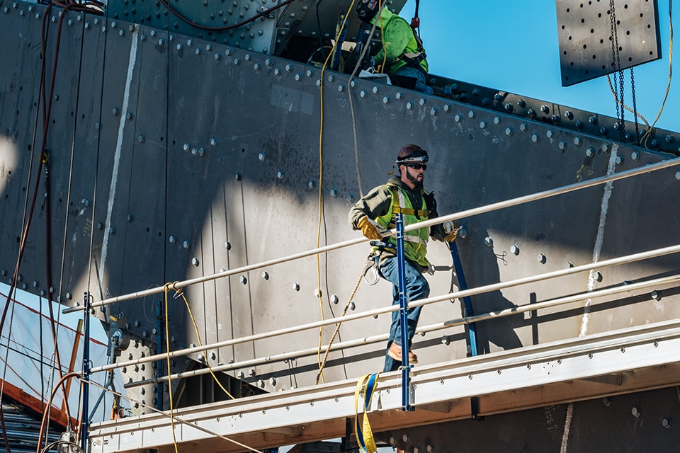 Construction worker wearing safety gear on a high platform, with metal structure in the background.