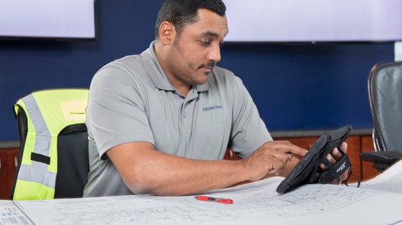 A person in a gray polo shirt works on a tablet over architectural blueprints in an office setting.