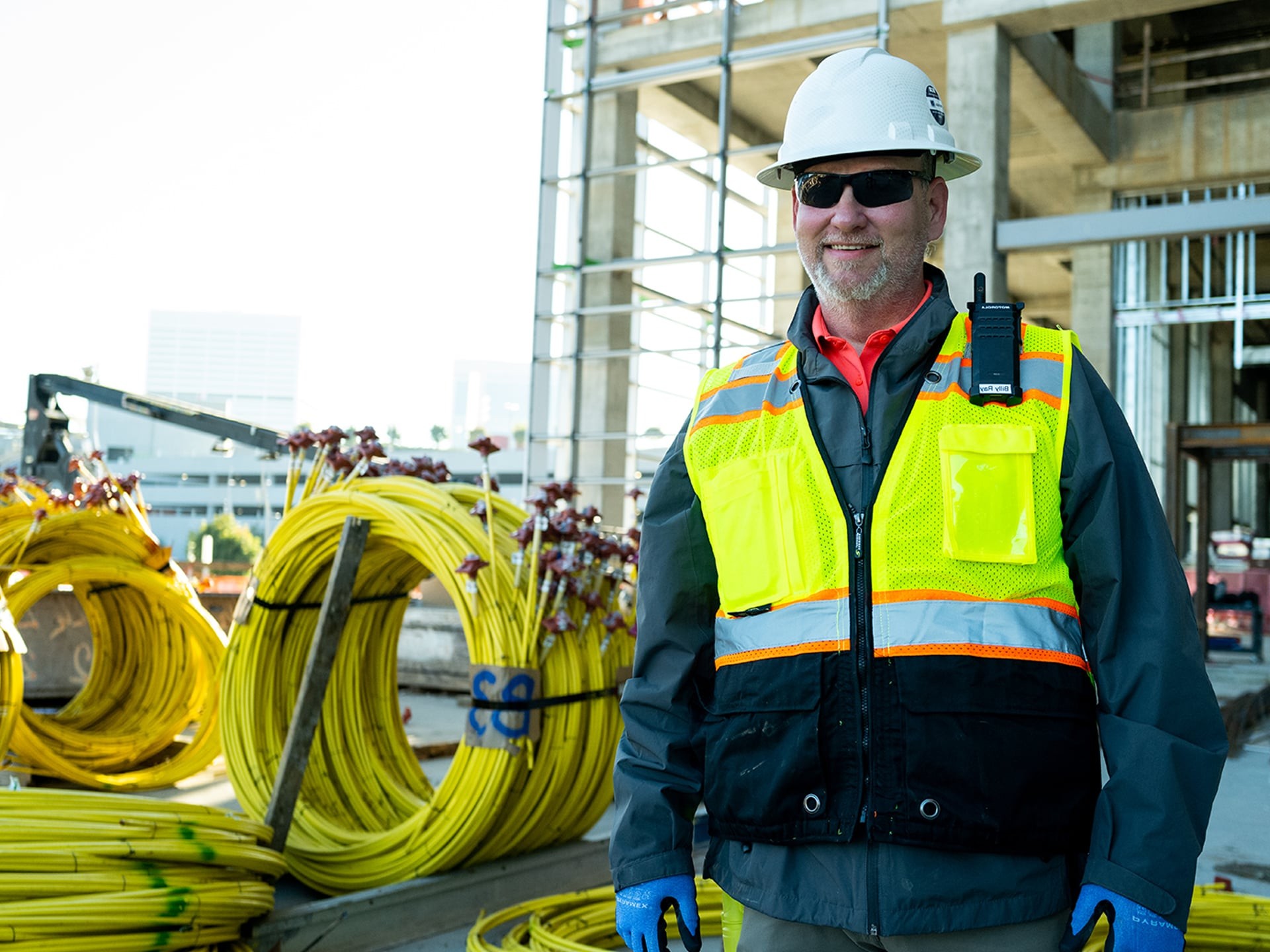 Construction worker with safety gear at building site with cables Construction worker in safety gear standing near coiled yellow cables at building site.