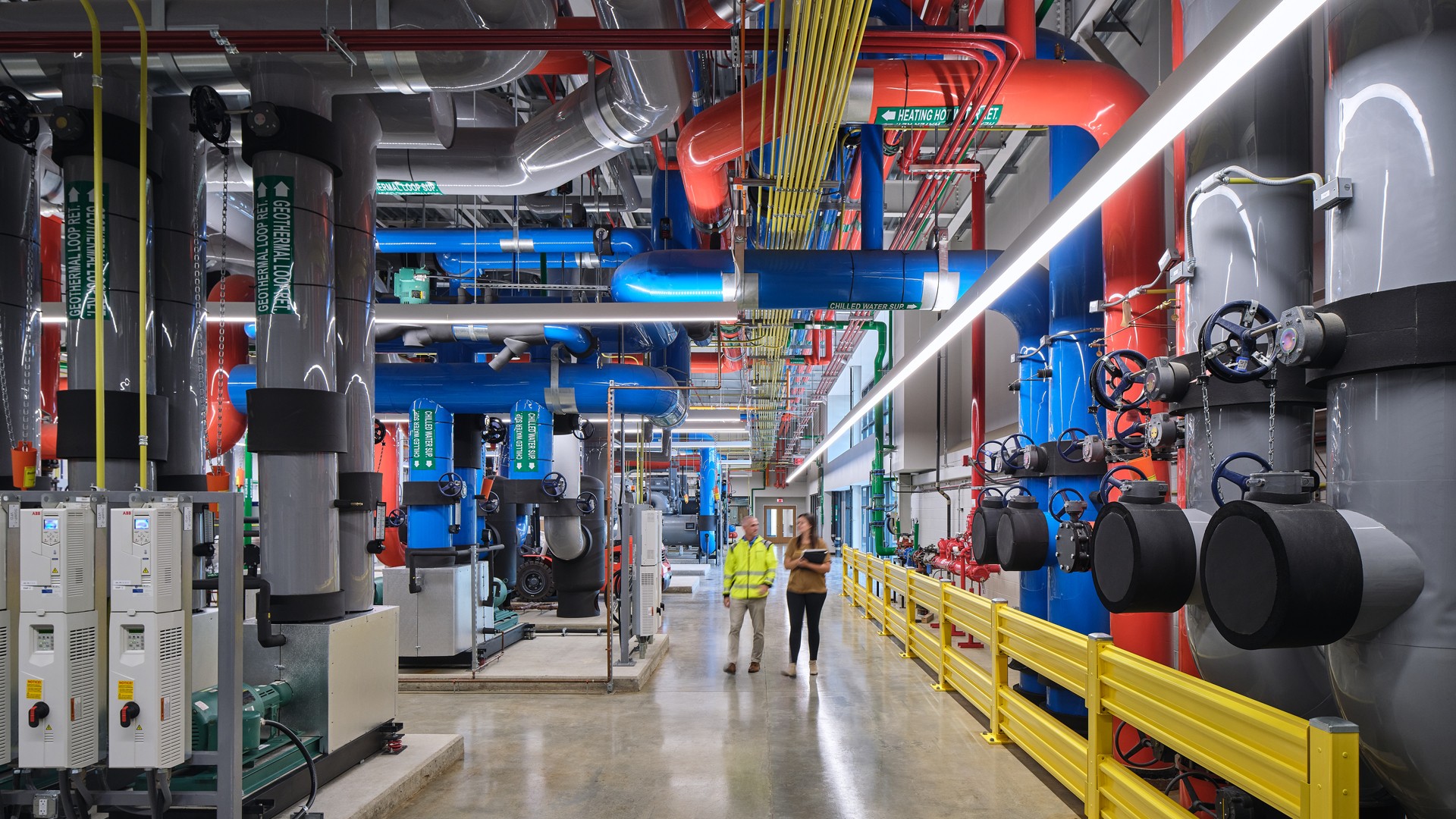 Industrial facility interior with complex piping and engineers Industrial facility interior with workers, colorful pipes, and safety railings.