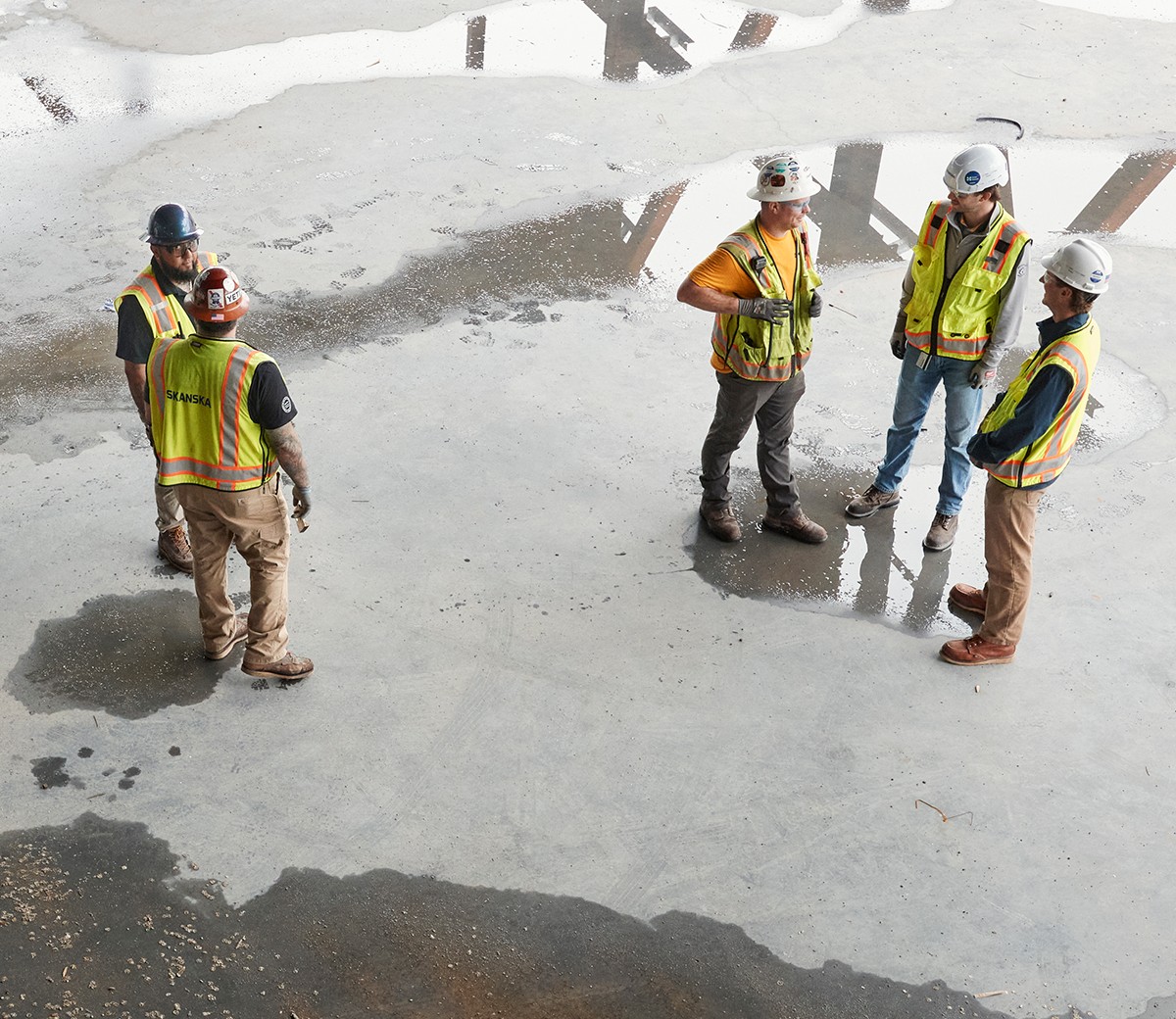 Construction workers in safety gear discussing on site Construction workers wearing safety vests and helmets standing on wet concrete floor.