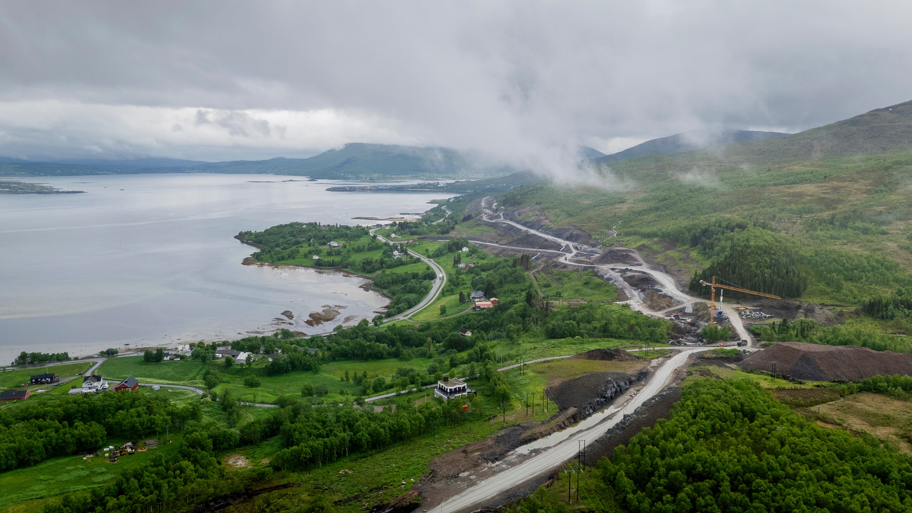 Luftfoto av en svingete vei gjennom frodig grønt landskap ved en rolig innsjø, under en skyet himmel.