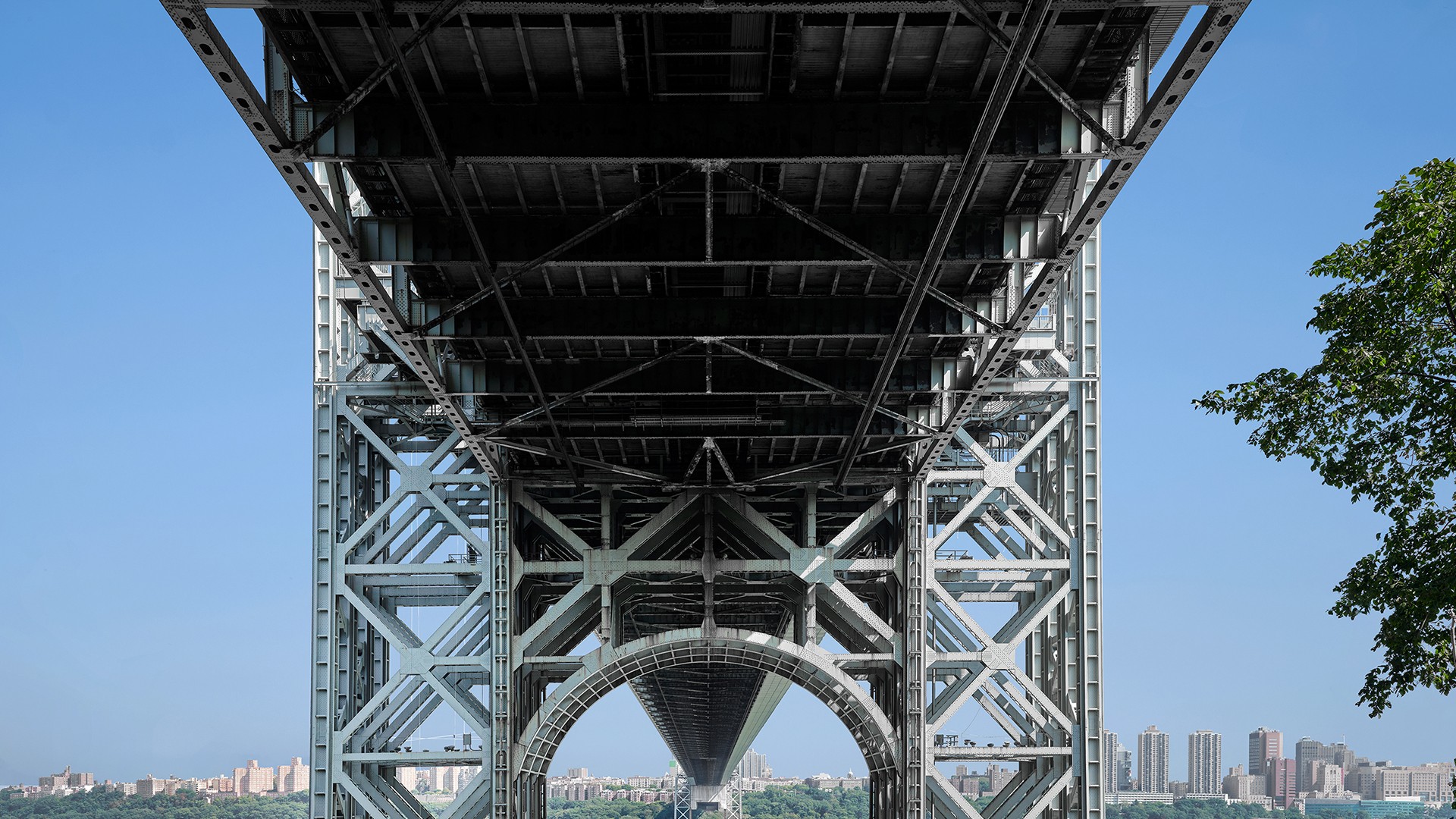Underside view of a steel bridge structure with city skyline and blue sky background.
