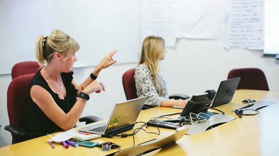 Two women collaborate at a conference table with laptops, discussing ideas on a whiteboard.
