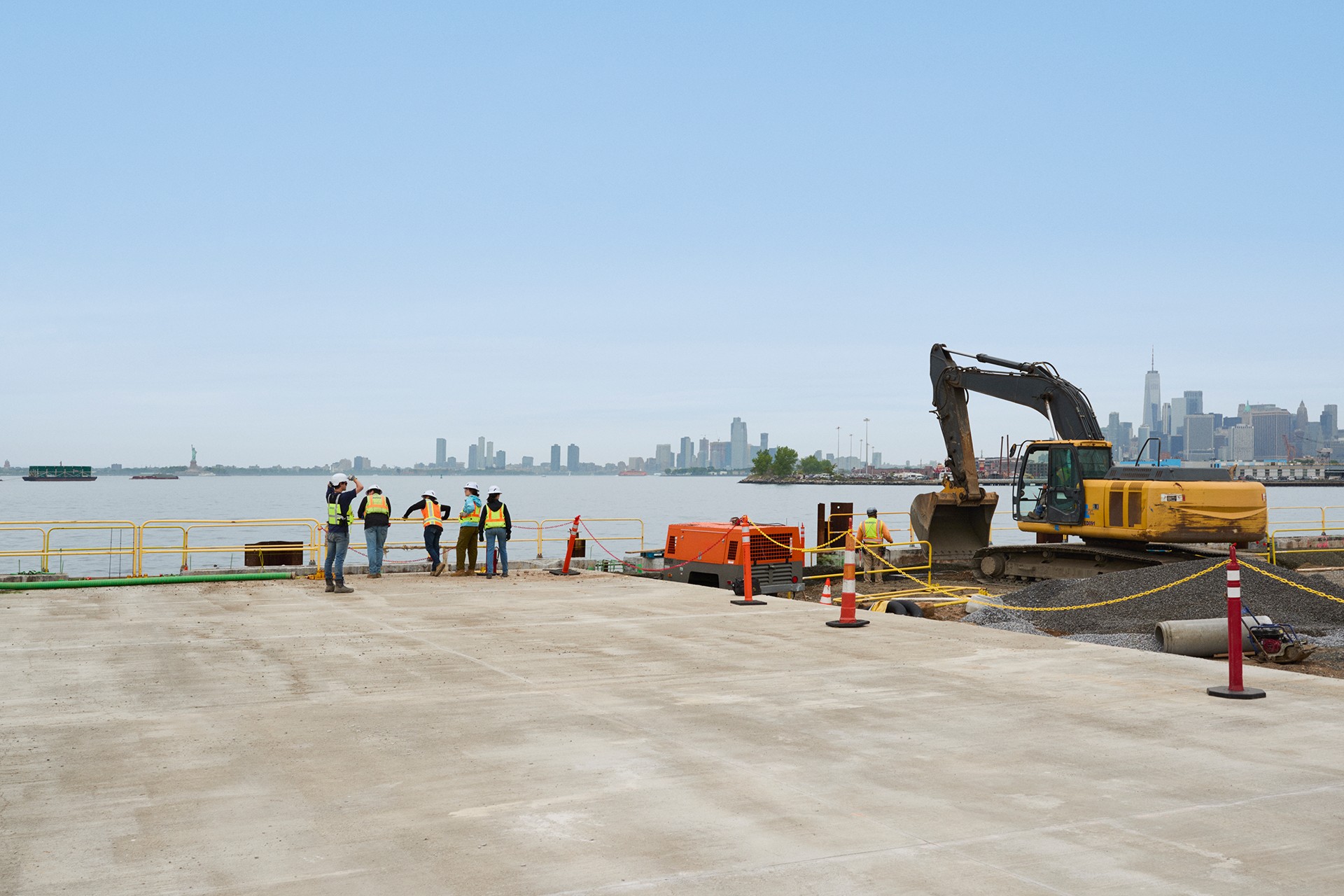 Construction workers and excavator at waterfront site with city skyline Construction workers and an excavator working near a waterfront with a city skyline view.