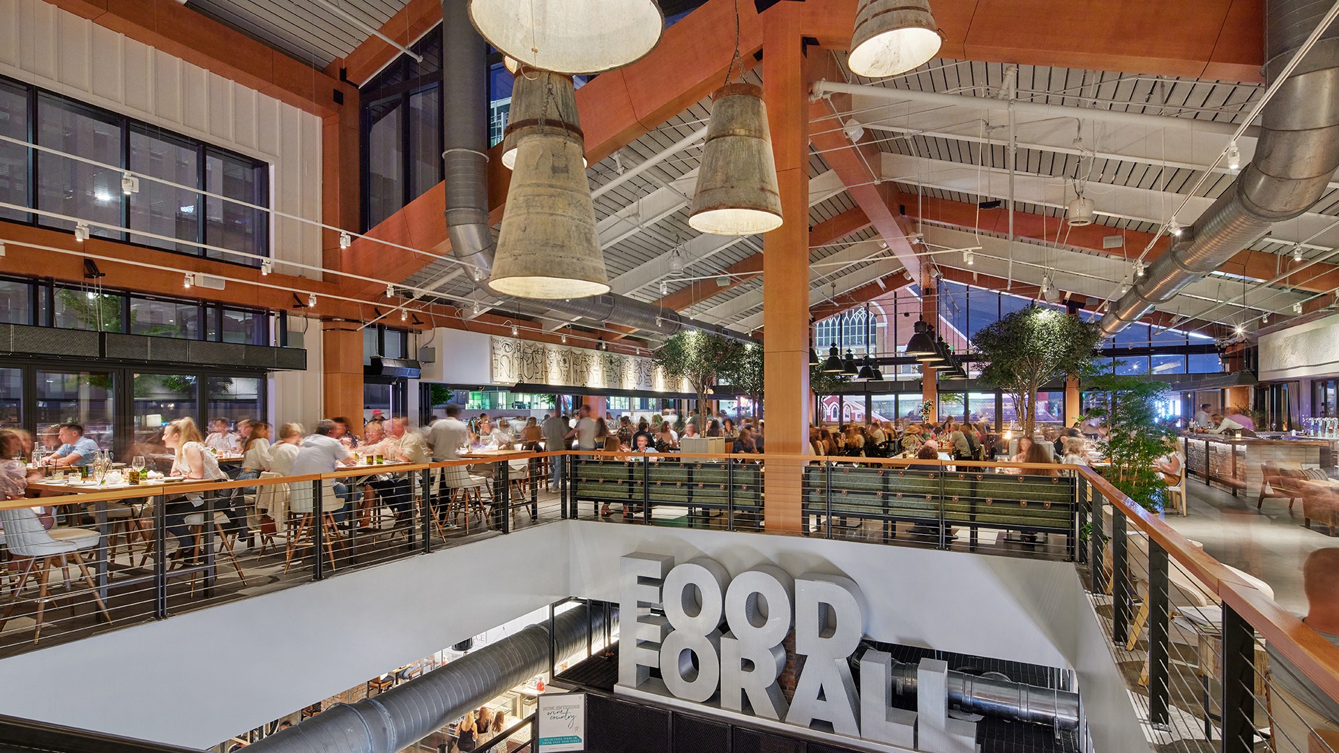 Vibrant food hall interior with bustling diners, large pendant lights, and "Food For All" sign.