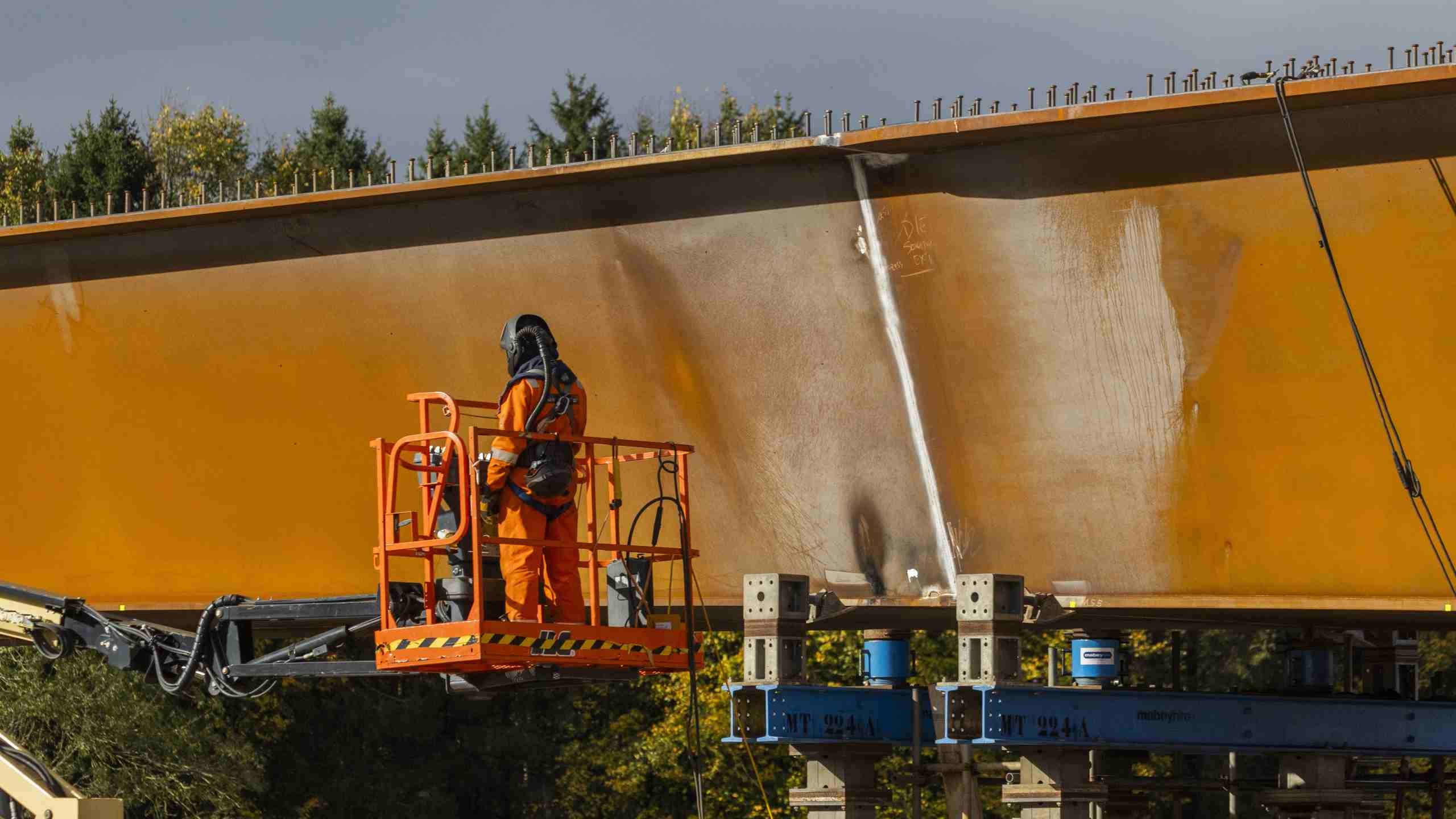 Bridge Construction Worker in Safety Gear on Lift Platform A worker in orange safety gear spray paints a large metal beam on a construction site platform.