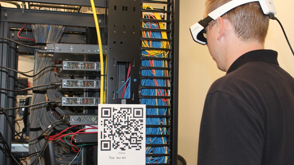 A person wearing AR glasses examines a server rack with colorful cables and a visible QR code.