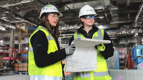 Two construction workers in safety gear review blueprints inside a building under construction.