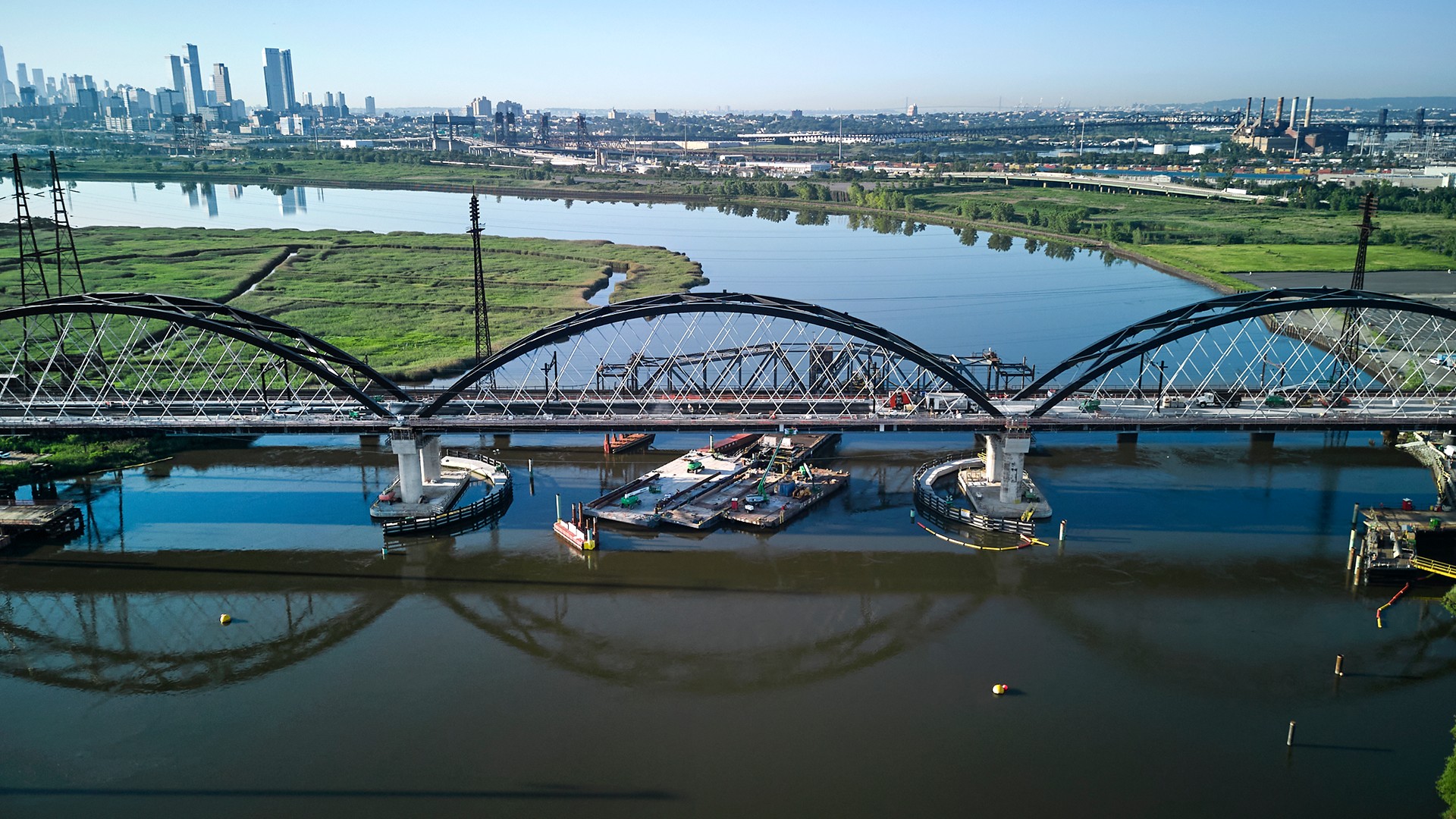 Aerial View of Arched Bridge Over Calm River with Cityscape Aerial view of a Portal North bridge over a river with city skyline and green wetlands.