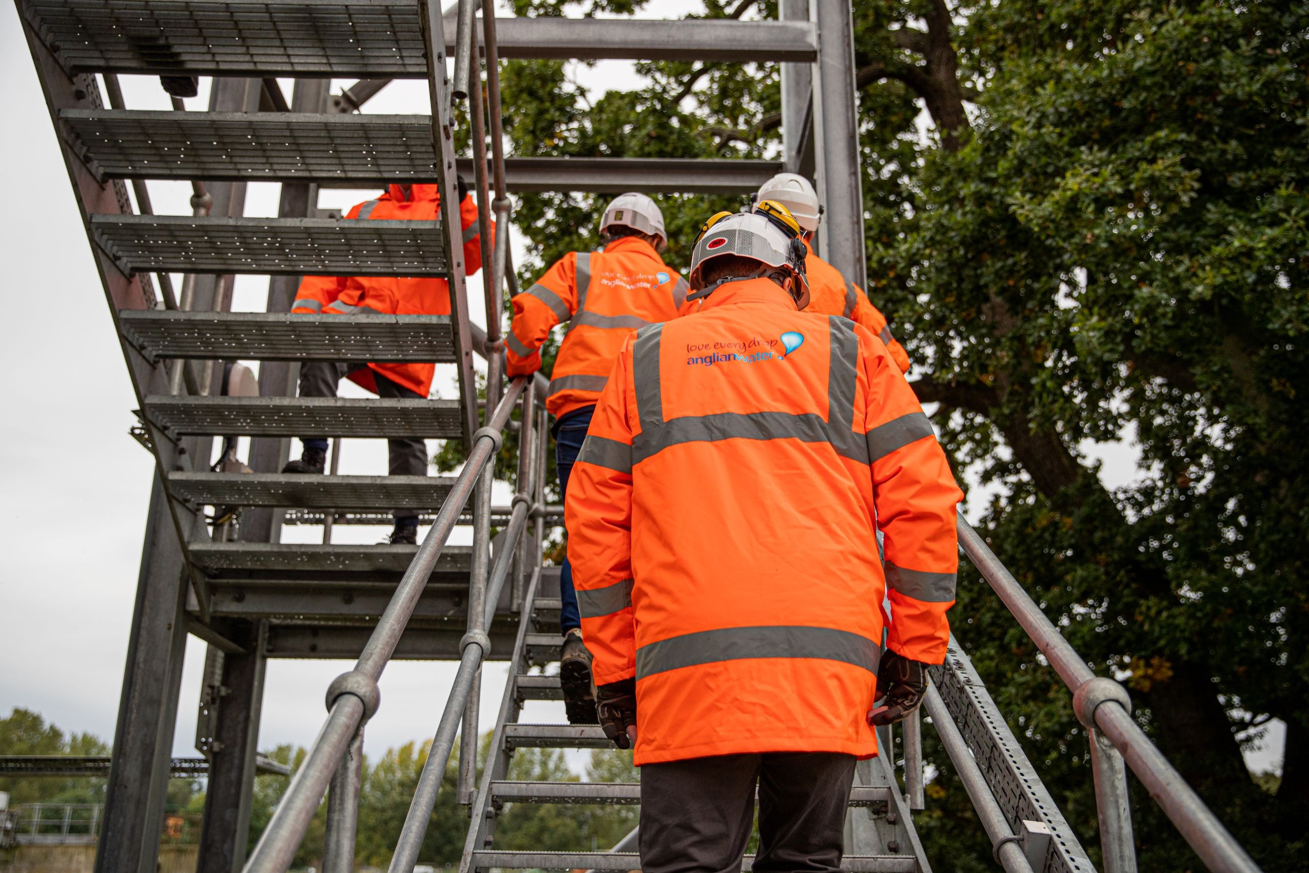 Workers in bright orange safety gear ascend a metal staircase at a construction site.