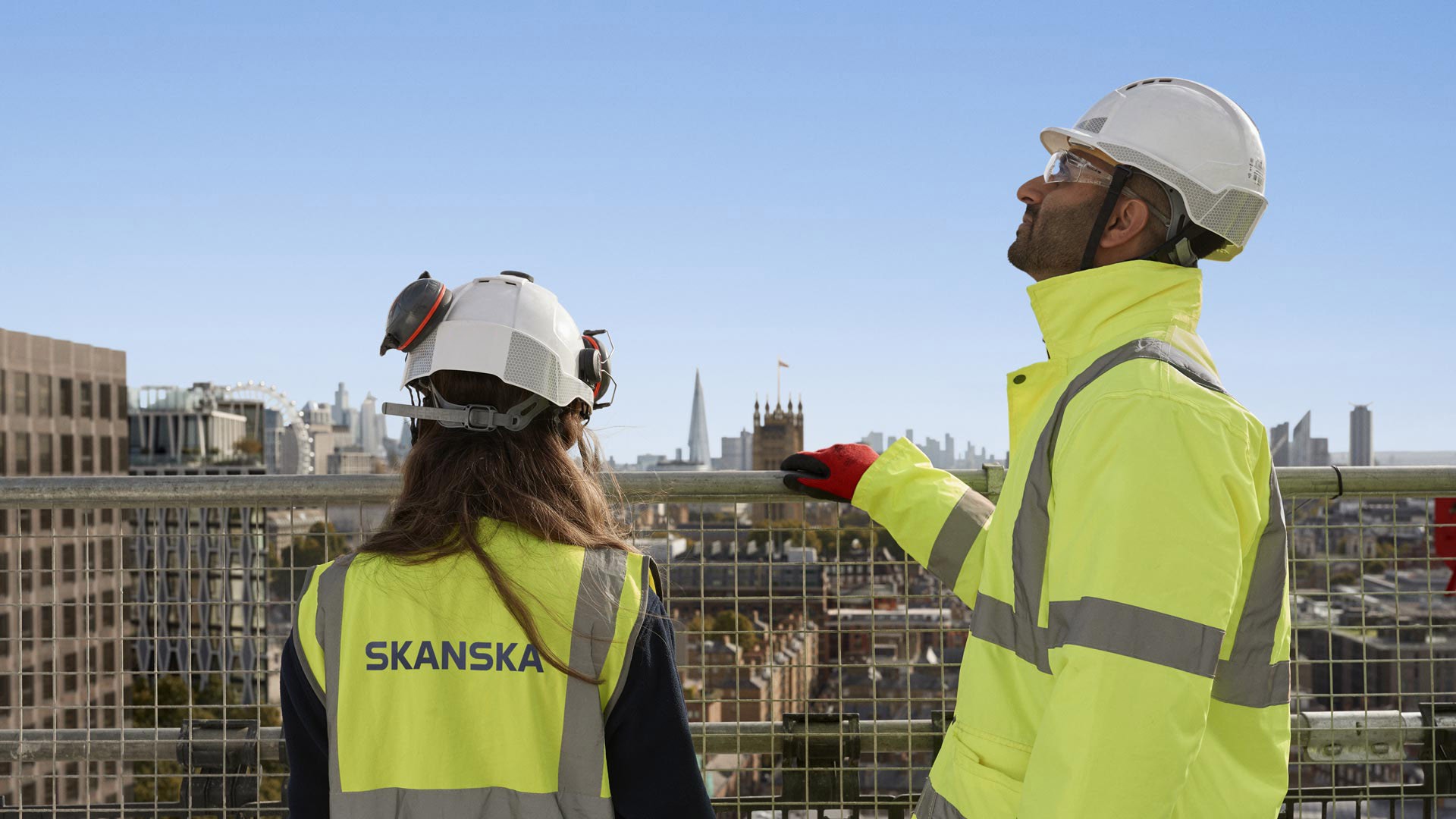 Construction Workers Overlooking London Skyline Two construction workers in Skanska safety gear overlook a cityscape from a high vantage point.