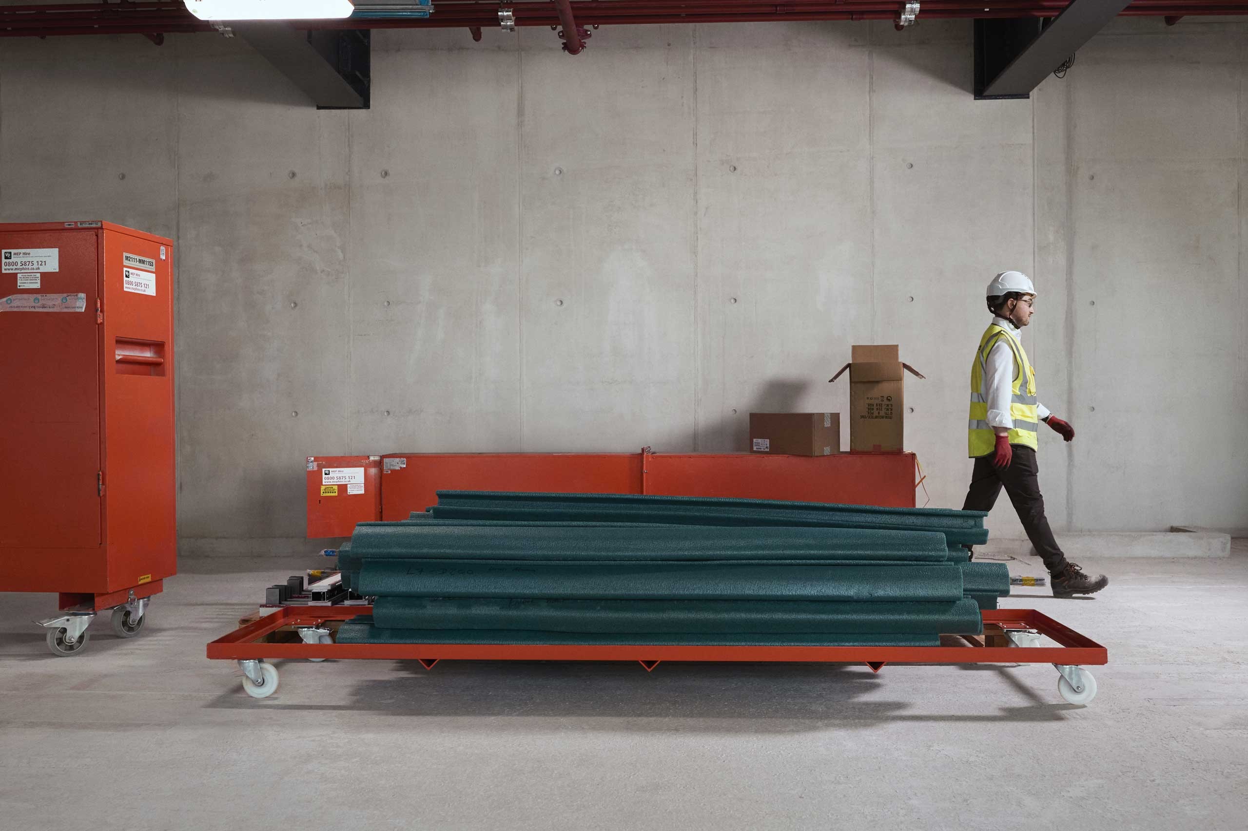 Construction worker in safety gear walking past stacked metal beams on a trolley indoors.